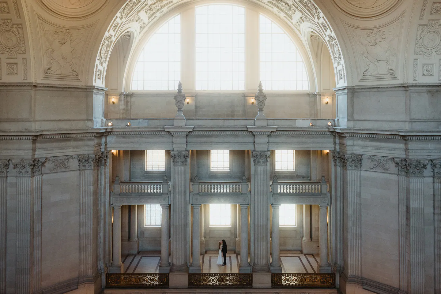 A newly married couple standing beneath the arched windows inside San Francisco City Hall