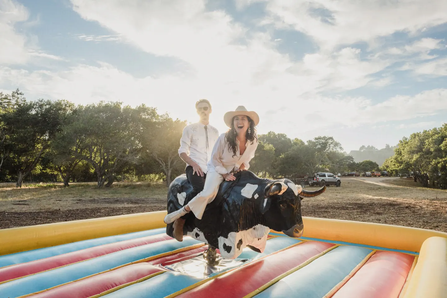 Newlyweds laughing together on a mechanical bull during their outdoor wedding