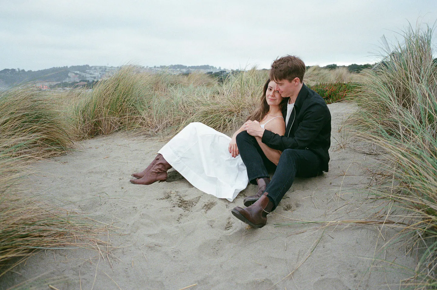 A newlywed couple sitting close together in coastal dunes, photographed on film